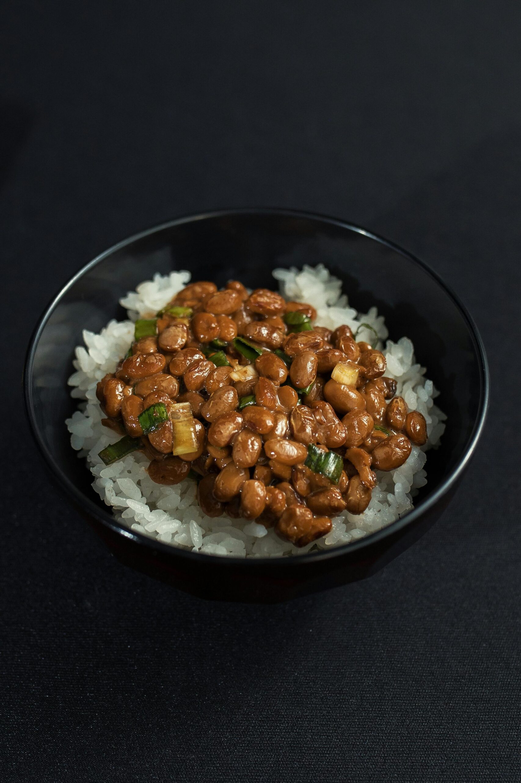 Appetizing natto served on steamed rice, garnished with green onions, in a sleek black bowl.